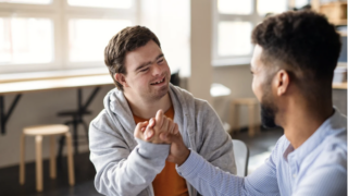 Two people sit indoors at a table, engaged in a friendly handshake. One person has Down syndrom and wears a gray hoodie, the other a striped shirt.